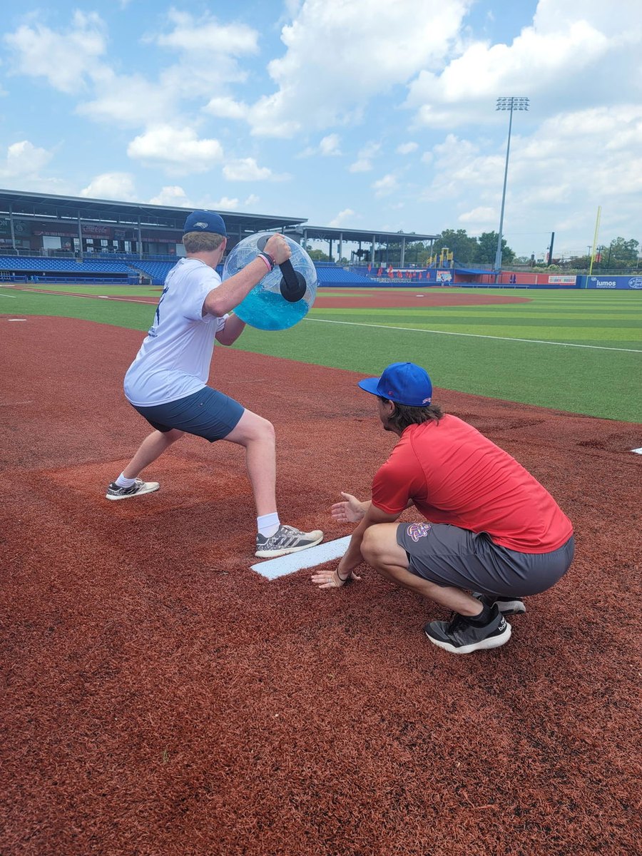 A youth player turning with a water-filled sphere on a baseball diamond while a coach watches.