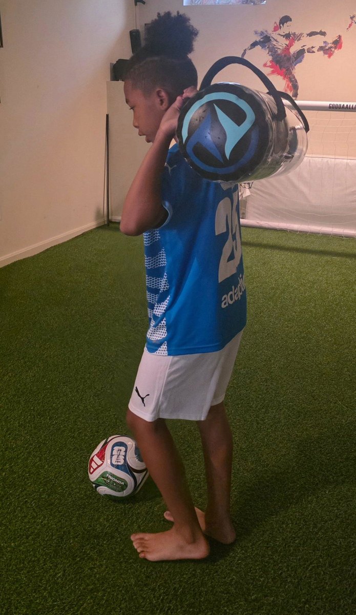 A young footballer on turf with a ball underfoot, carrying a water-filled sphere.